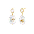 Gold-plated drop earrings with iridescent baroque pearls and crystal flower accents on cream background